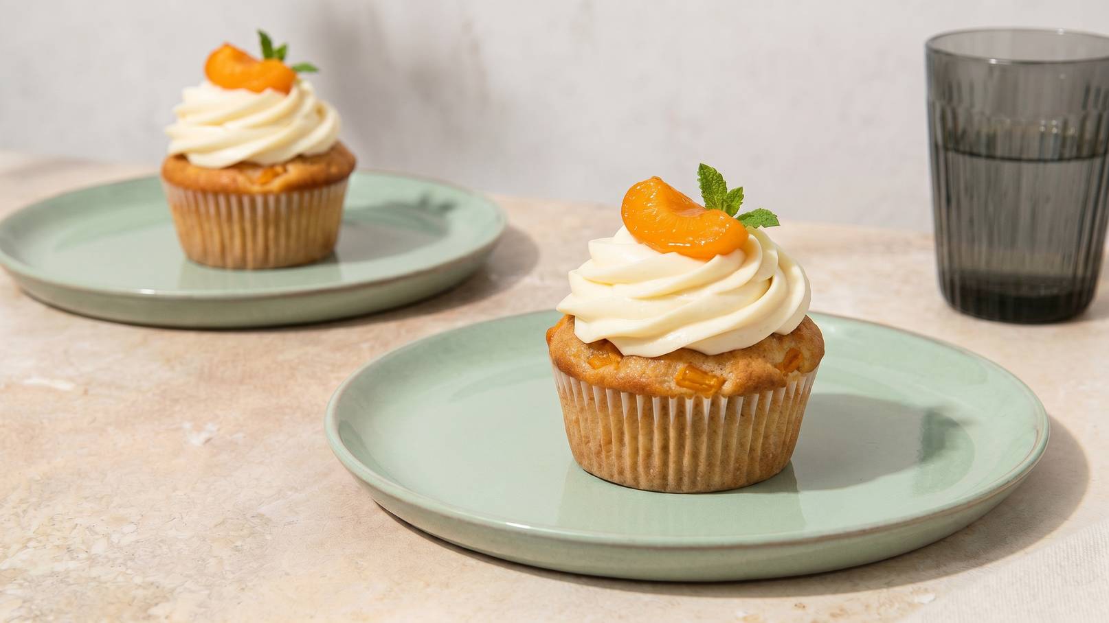 Auf dem Bild sind zwei Cupcakes auf flachen, grünen Tellern zu sehen. Jeder Cupcake ist mit einer großzügigen Menge weißer Sahne dekoriert und mit einer Halbe Mandeline und einem Minzblatt garniert. Im Hintergrund steht ein schwarzes Glas.