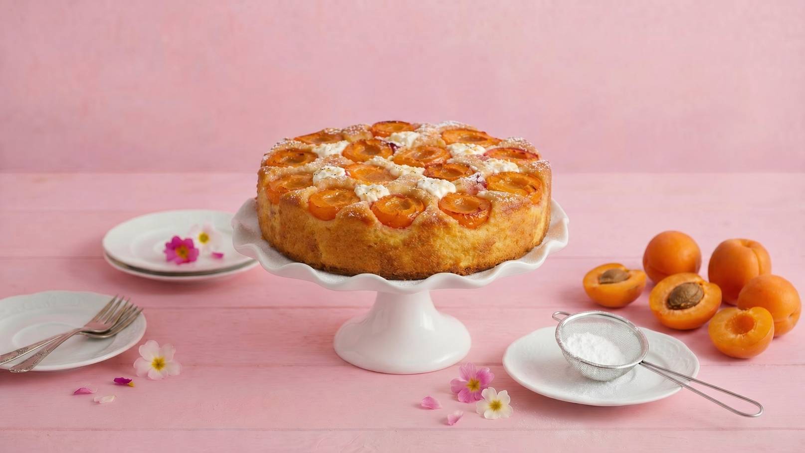 Ein geflochtenes Aprikosenkuchen steht auf einem weißen Tortenständer. Der Kuchen ist mit Aprikosenhalben garniert und mit Puderzucker bestäubt. Daneben liegen frische Aprikosen und kleine Teller mit Besteck. Der Hintergrund ist zart rosa.