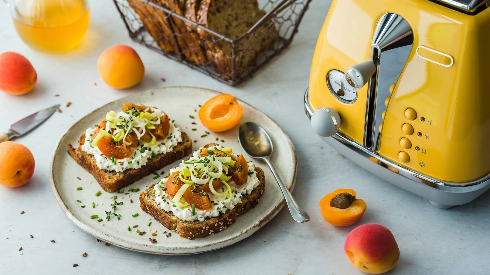 Auf dem Teller liegen zwei Scheiben Vollkornbrot, belegt mit Quark, frischen Aprikosen und geschnittenem Lauch. Daneben liegt ein Löffel. Im Hintergrund steht ein gelber Toaster und eine Schale mit weiteren Aprikosen.