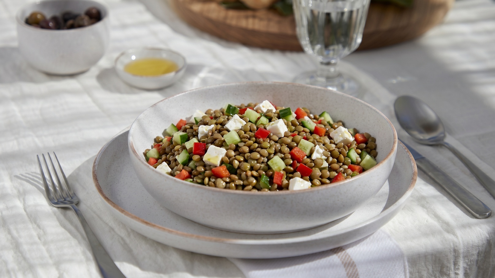 Eine Schüssel mit Linsensalat steht auf einem Tisch. Der Salat enthält grüne Linsen, gewürfelte Paprika, Gurken und kleine Stücke Feta-Käse. Die Präsentation ist frisch und ansprechend, umgeben von einem Essbesteck und weiteren Schalen im Hintergrund.