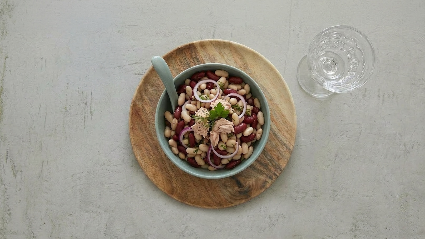 Eine Schüssel mit buntem Bohnensalat, in der sich rote und weiße Bohnen sowie Zwiebelringe und Stücke von Thunfisch befinden. Die Schüssel ist auf einem runden Holzbrett platziert, daneben steht ein Glas Wasser.