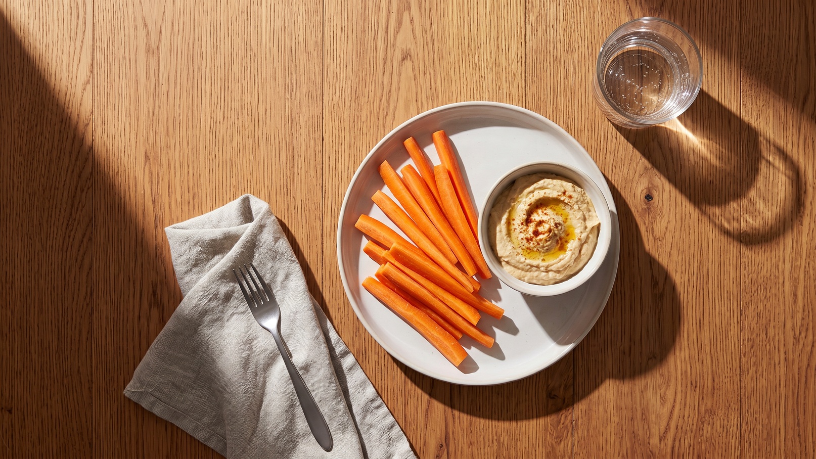 Ein Teller mit frischen Karottensticks liegt neben einer kleinen Schüssel mit Hummus. Daneben befindet sich ein Besteckset und ein Glas Wasser auf einem Holztisch. Die Szene ist hell erleuchtet und vermittelt einen gesunden Snack.