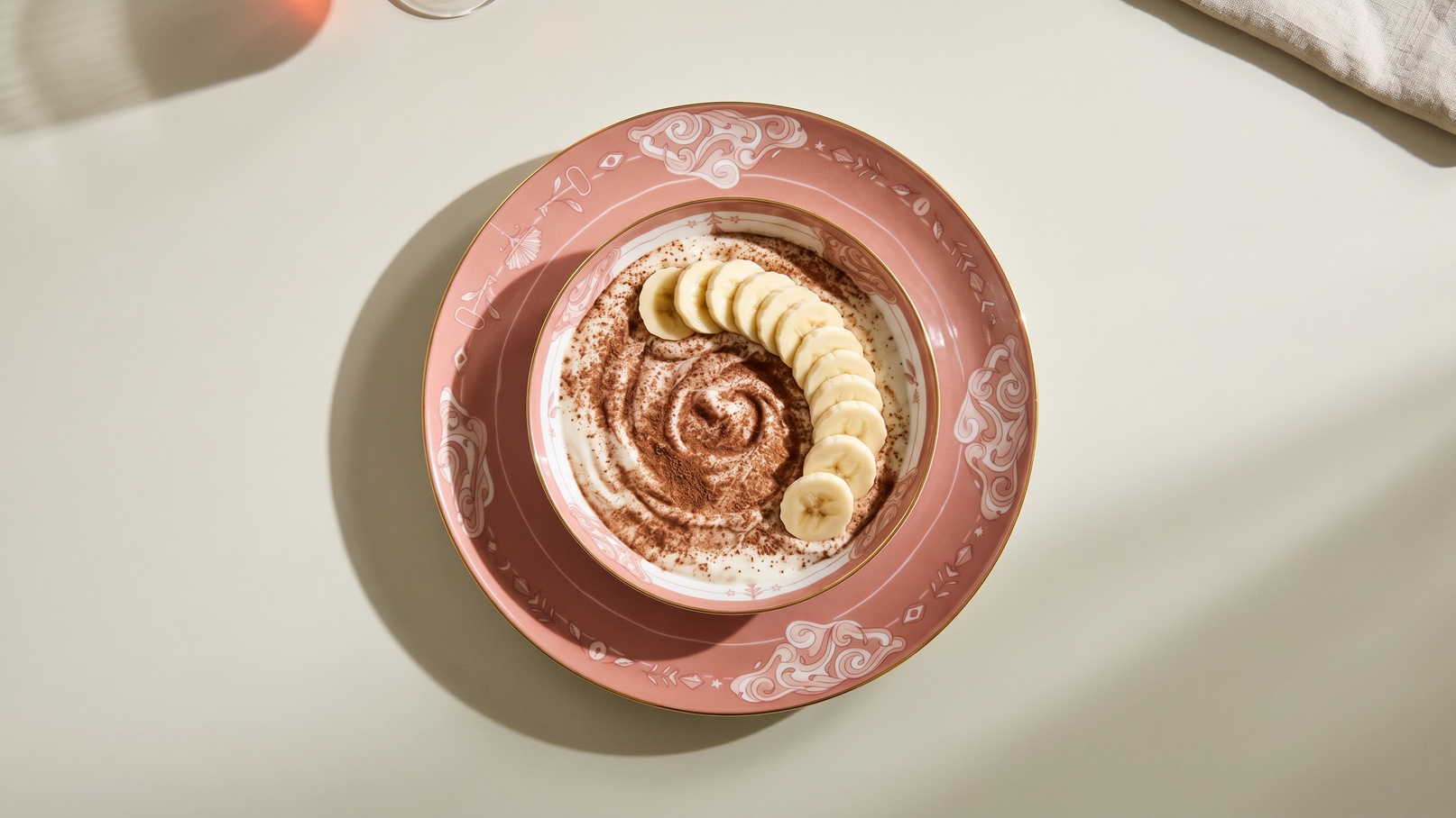 Eine rosa Schüssel mit einer cremigen Schicht, dekoriert mit Kakaopulver und Banane in Scheiben, die spiralartig angeordnet sind. Die Präsentation des Desserts gibt ihm ein ansprechendes Aussehen.