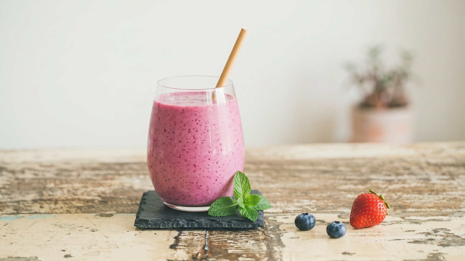Ein Glas mit einem lila Smoothie auf einem schwarzen Untersetzer. Der Smoothie hat einen Strohhalm und ist mit einem Minzblatt garniert. Neben dem Glas liegen eine Erdbeere und einige Heidelbeeren auf einem rustikalen Holztisch.
