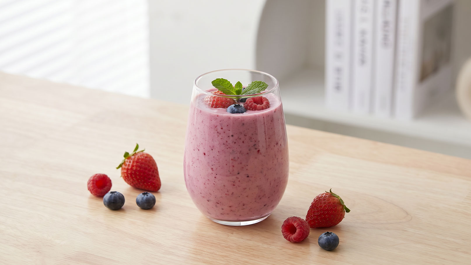 Ein Glas mit einem pinkfarbenen Smoothie, garniert mit Himbeeren und einem Minzblatt, steht auf einem Holztisch. Um das Glas herum liegen frische Erdbeeren, Himbeeren und Heidelbeeren.