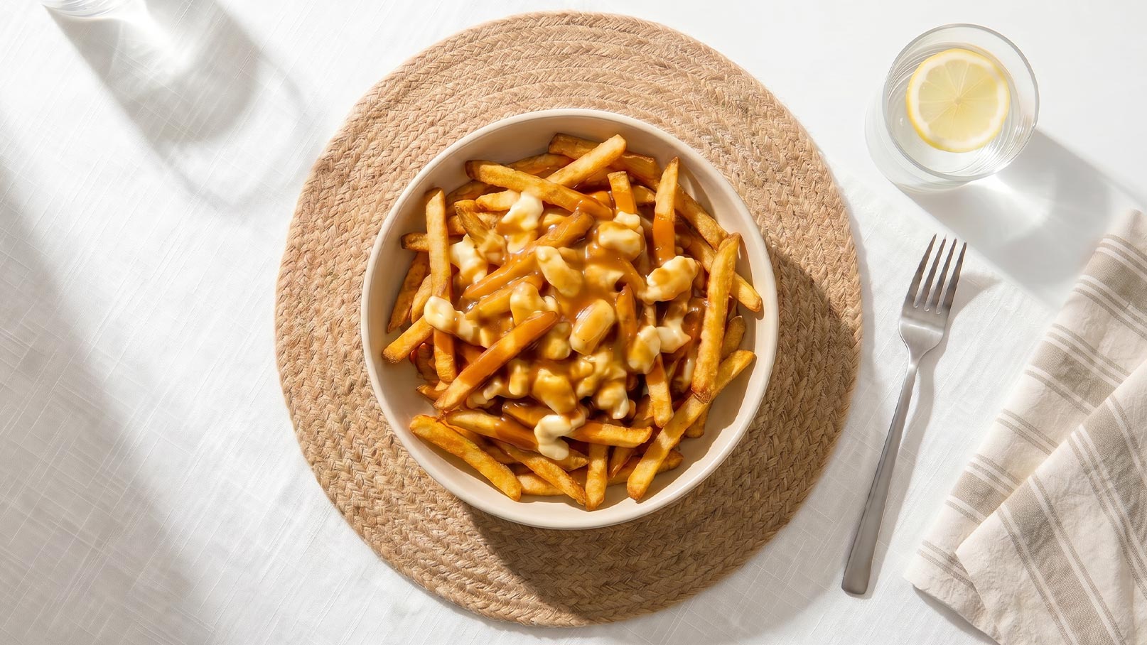 Eine Schüssel mit Pommes Frites, überzogen mit einer cremigen Käsesauce, steht auf einem runden Untersetzer. Daneben liegt ein Besteck und ein Glas Wasser mit einer Zitronenscheibe. Der Tisch ist schlicht und hell.