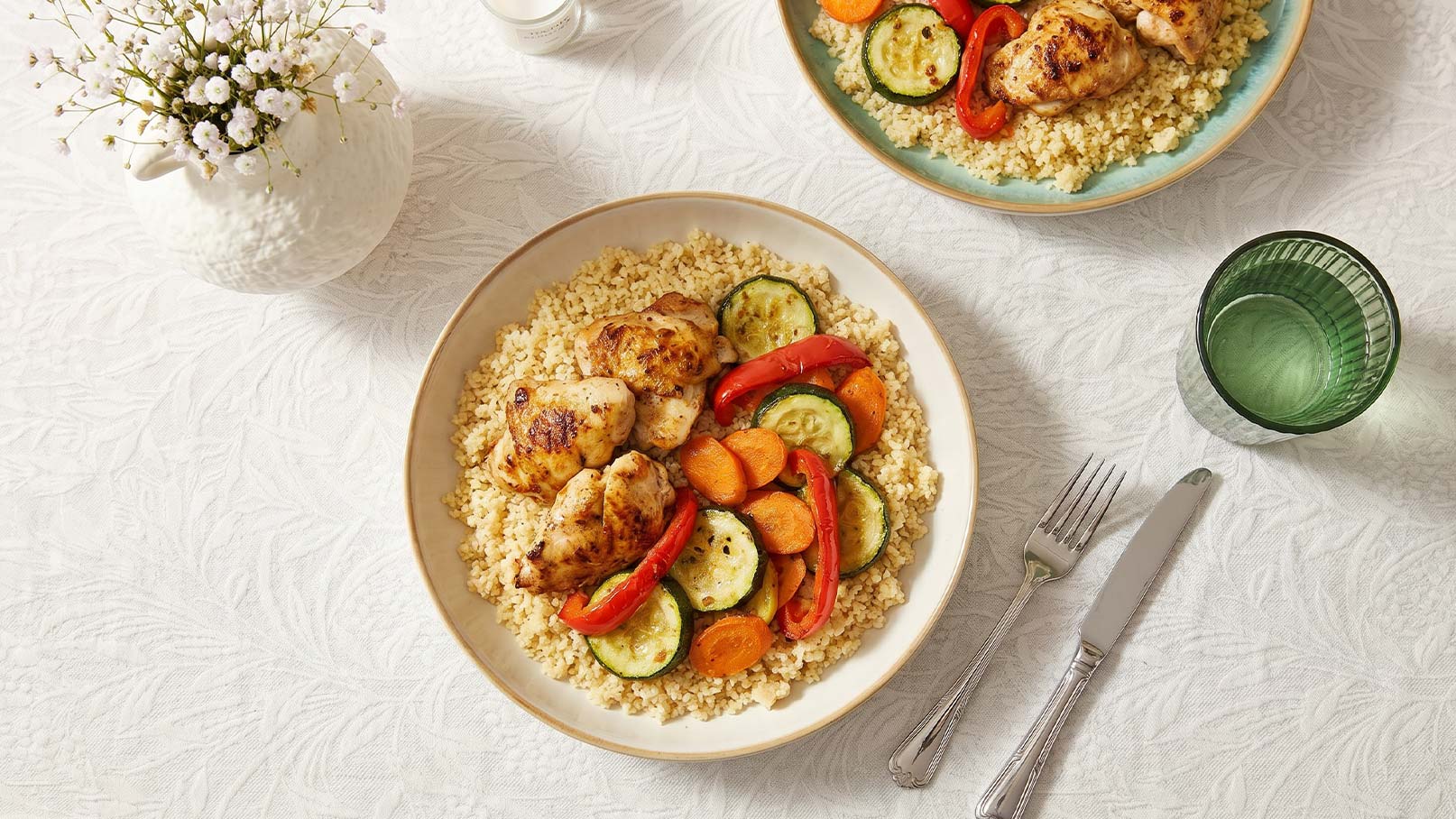 Ein Teller mit Couscous, gegrilltem Hähnchen und einer bunten Mischung aus Gemüse wie Zucchini, Karotten und Paprika. Der Tisch ist schlicht gedeckt, mit einem Glas und einer kleinen Blumenvase im Hintergrund.