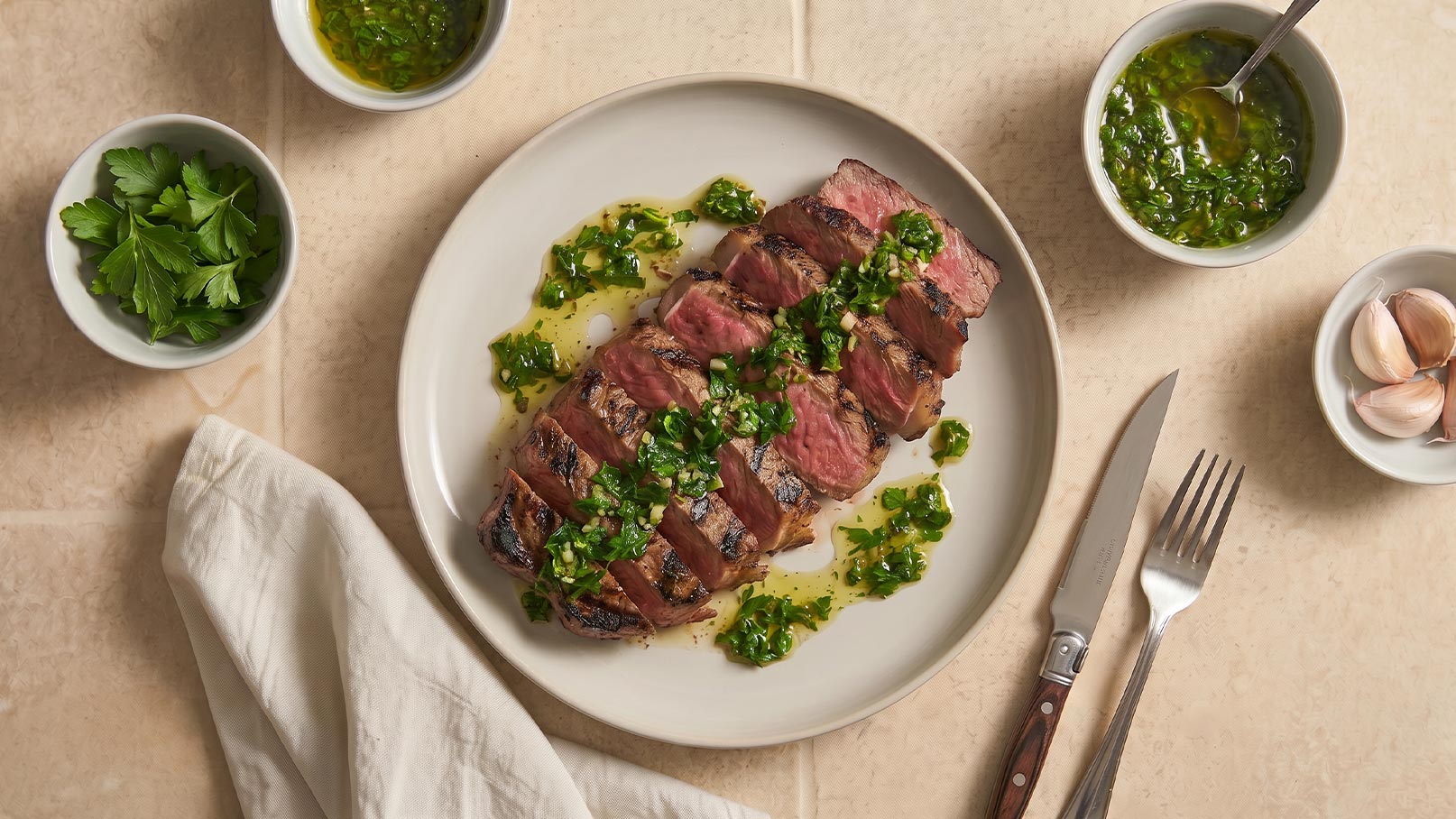 Auf dem Bild liegt ein aufgeschnittener Steak auf einem Teller, umgeben von grüner Sauce und frischen Kräutern. Neben dem Teller stehen mehrere kleine Schalen mit einer ähnlichen Sauce und Kräutern sowie einer Knoblauchknolle. Ein Besteckset liegt bereit.