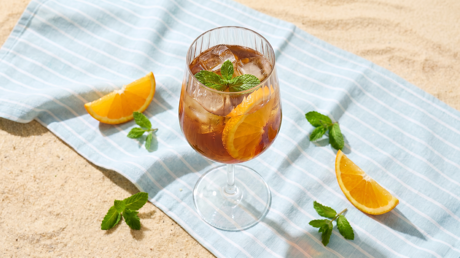 Ein Glast mit einem erfrischenden Getränk, gefüllt mit Eis, Orangenscheiben und einem Minzblatt. Das Glas steht auf einem gestreiften Tuch, umgeben von weiteren Orangenscheiben und frischen Minzblättern, vor einem sandigen Hintergrund.