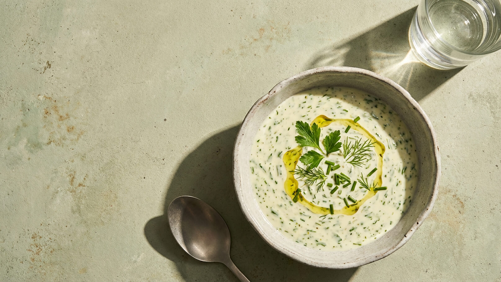 Ein tiefes Schüsselgericht mit einer cremigen, hellgrünen Suppe. Die Oberfläche ist mit frischen Kräutern wie Petersilie und Dill sowie einem Spritzer Olivenöl garniert. Daneben liegt ein Löffel auf einer matten Oberfläche, und im Hintergrund steht ein Glas Wasser.