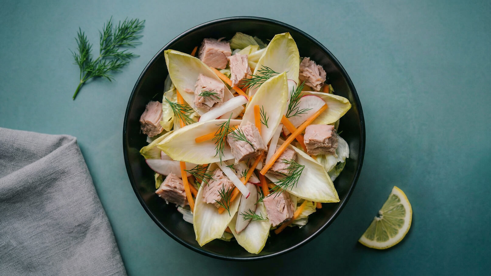 Eine Schüssel mit Salat, bestehend aus Endiviensalat, Thunfischstücken, Karottenstreifen und Radieschen. Frische Dillzweige sind darüber verteilt. Ein Zitronenschnitz liegt daneben auf der Tischoberfläche. Der Hintergrund ist in einer sanften grünen Farbe gehalten.