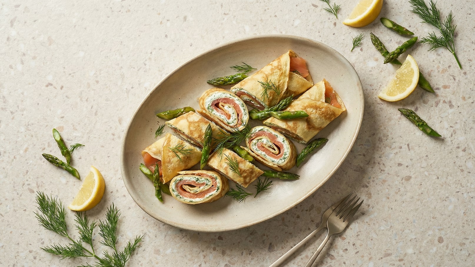 Auf einem ovalen Teller sind geschnittene Pfannkuchen-Rollen gefüllt mit Lachs und Kräutercreme angerichtet. Neben den Rollen liegen frische grüne Spargelstücke und einige Zitronenscheiben, die dem Gericht eine frische Note verleihen.
