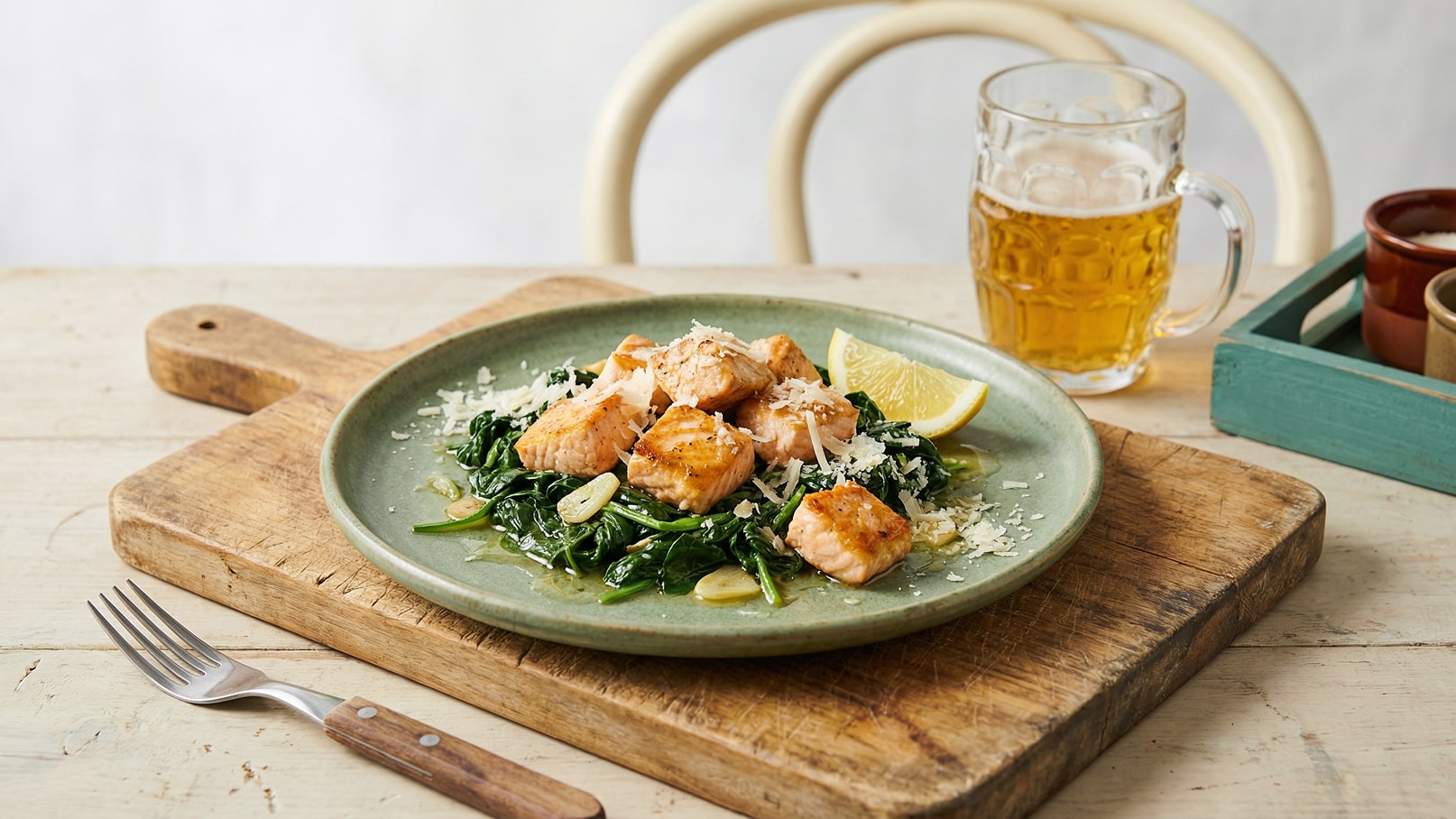 Ein Teller mit gebratenem Lachs auf einem Bett aus Spinat, garniert mit Zitronenscheiben und Käse. Der Teller steht auf einem Holzbrett, daneben liegt eine Gabel. Im Hintergrund ist ein Glas Bier zu sehen.