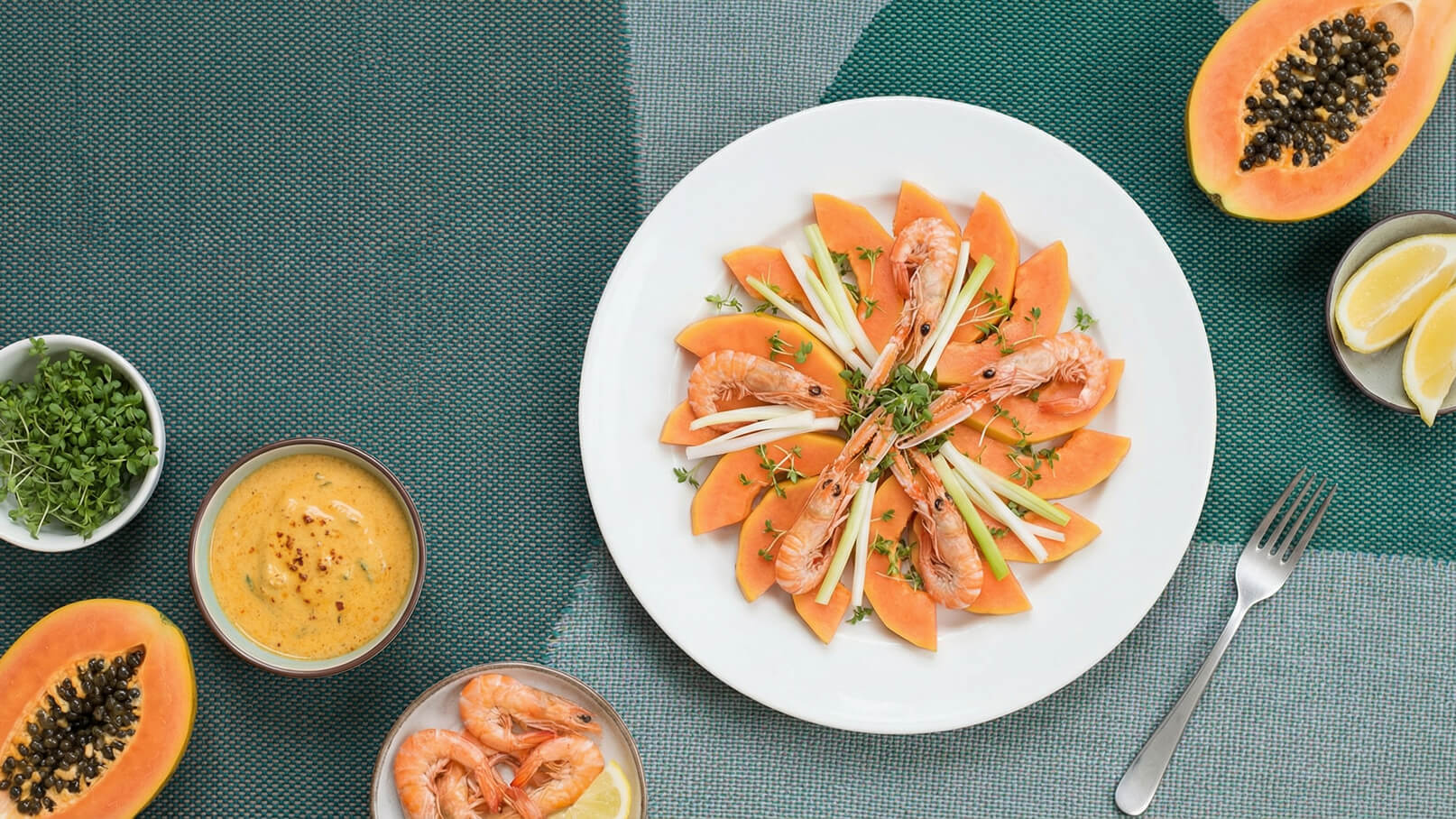 Ein Teller mit frischer Papaya, geschält und in Spalten geschnitten, dekoriert mit gekochten Garnelen und frischem Schnittlauch. Daneben eine Schale mit einer cremigen Sauce und weitere Garnelen. Im Hintergrund liegen Hälften einer Papaya und Zitronenschnitze.