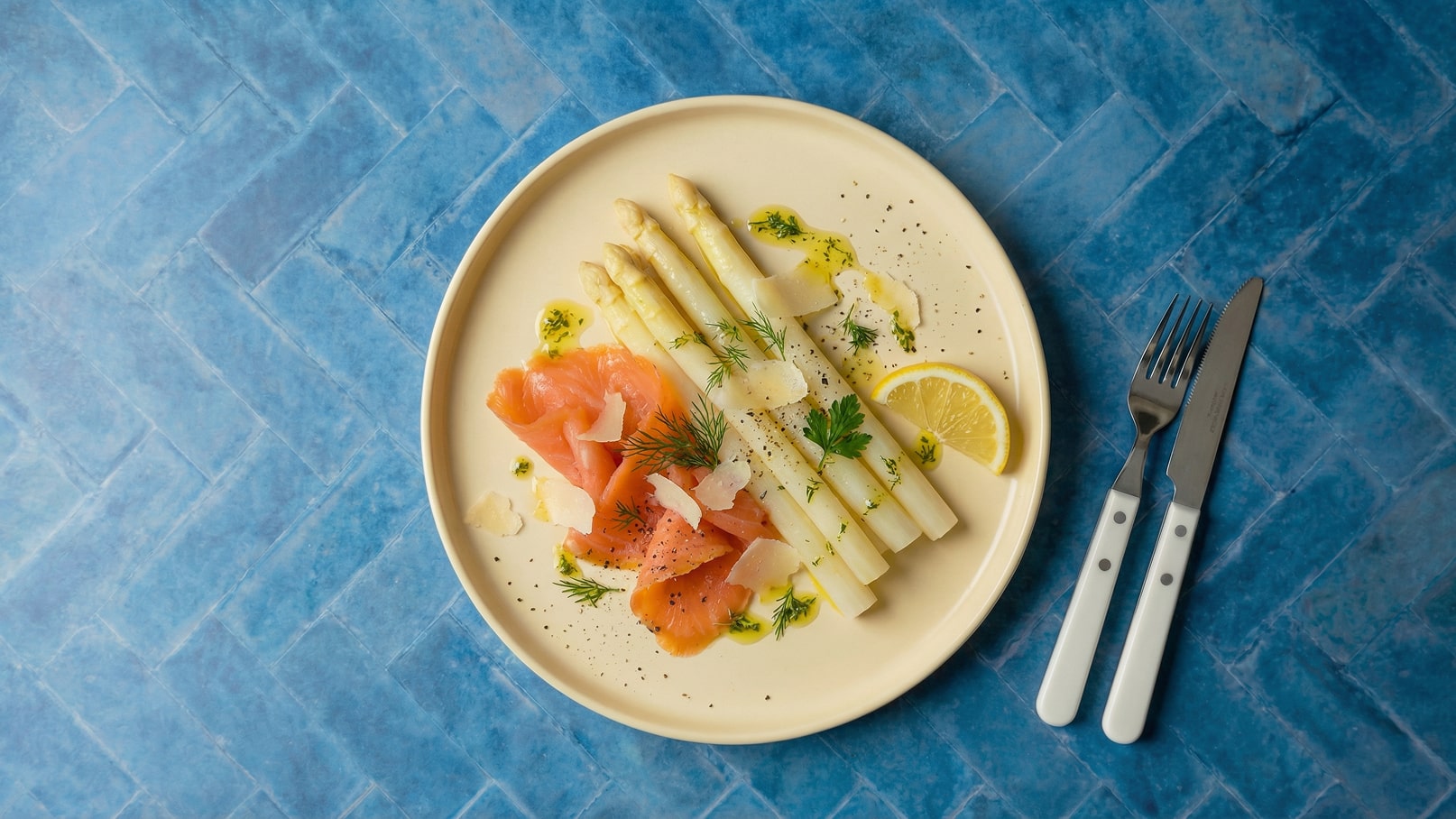 Ein Teller mit weißen Spargelstangen liegt auf einer blauen Unterlage. Daneben befinden sich Räucherlachs, frische Kräuter und ein Stück Zitrone. Das Gericht ist mit Olivenöl und Parmesanspänen garniert, alles schön angerichtet und appetitlich.
