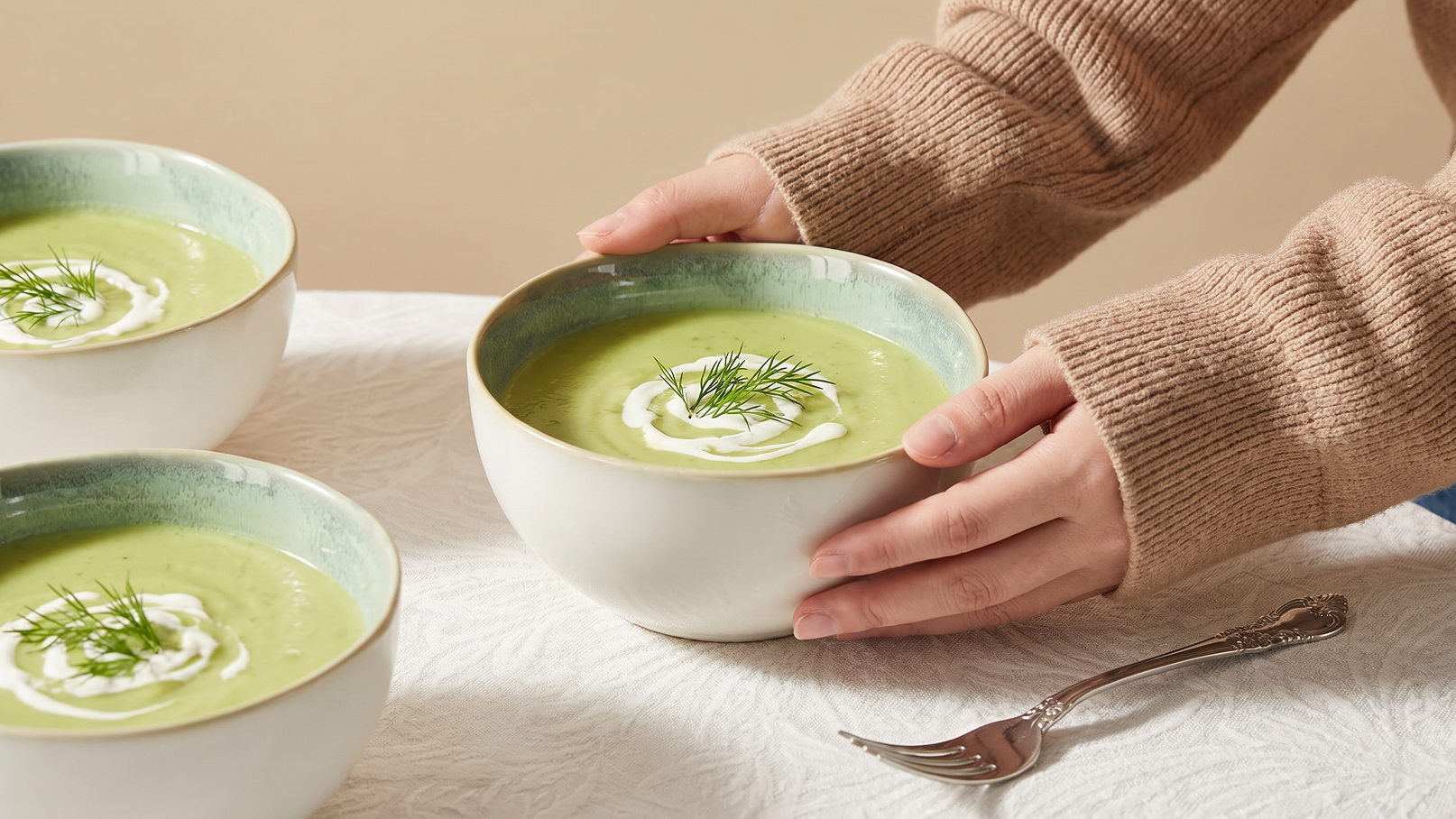 Eine Person mit beigem Pullover hebt eine Schüssel mit grüner Suppe, die mit Sahne und frischem Dill garniert ist. Im Hintergrund stehen weitere Schalen mit derselben Suppe. Der Tisch hat eine helle, strukturierte Tischdecke.