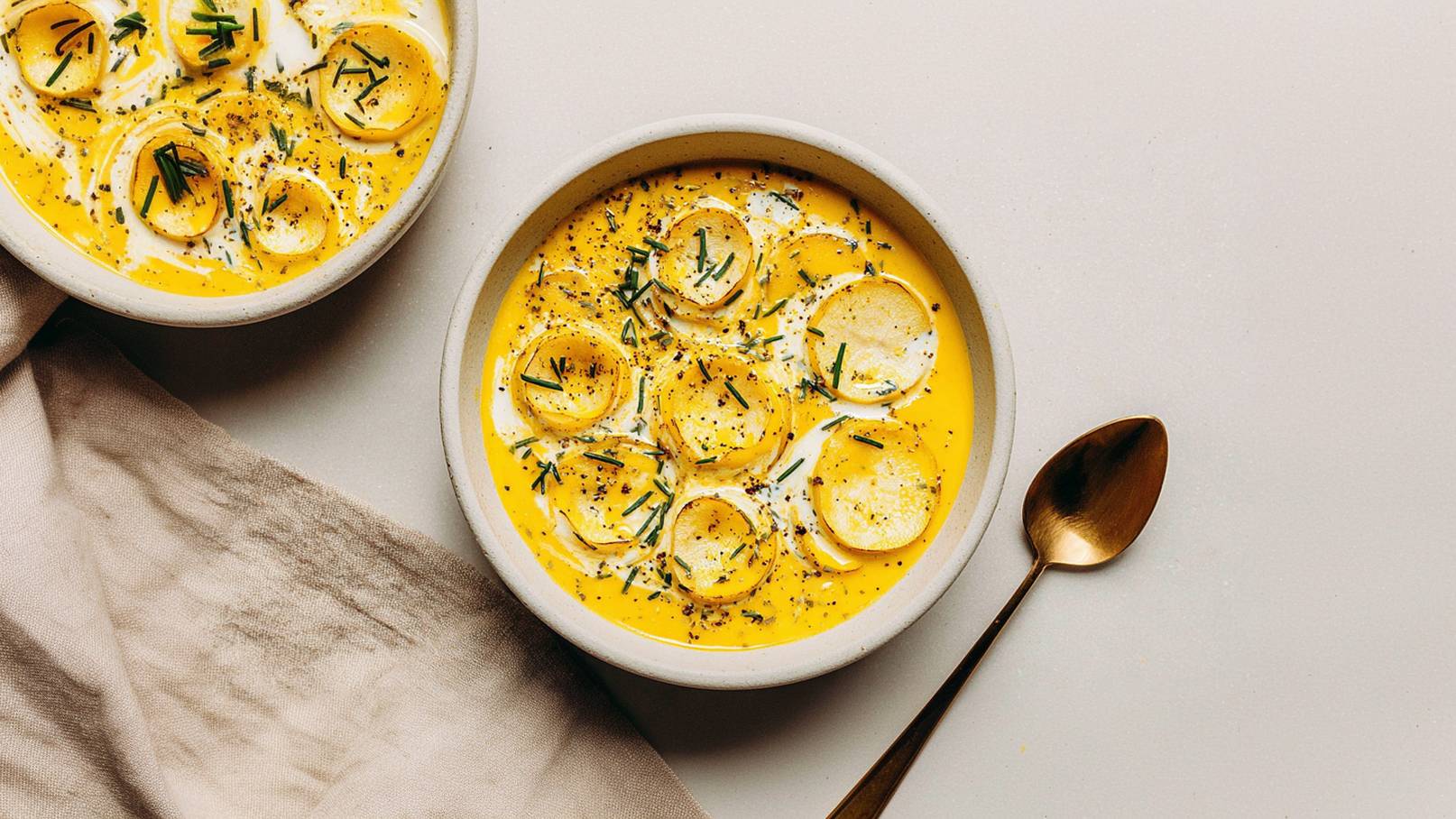 Eine Schüssel mit cremiger, gelber Suppe, garniert mit runden, weißen Scheiben und frischem Schnittlauch. Neben der Schüssel liegt ein goldener Esslöffel. Der Hintergrund ist hell und schlicht.