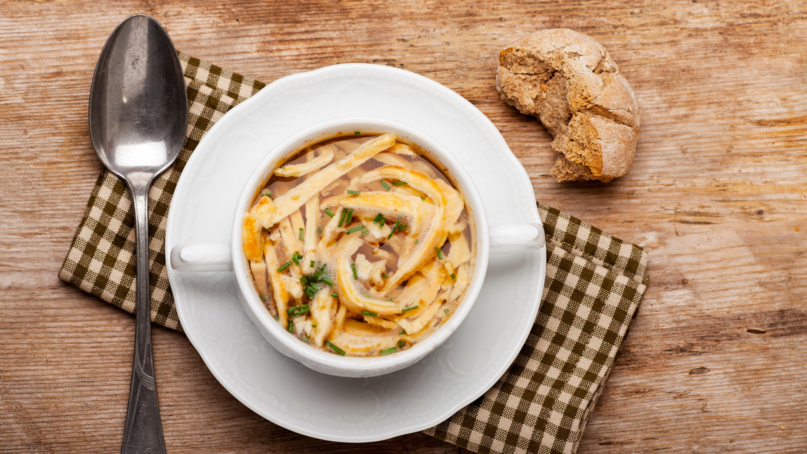 Eine Schüssel mit klarer Brühe, gefüllt mit feinen Nudeln und frischem Schnittlauch. Daneben liegt ein Stück dunkles Brot auf einem karierten Stoffuntersetzer. Eine silberne Löffel ist ebenfalls zu sehen, auf einem rustikalen Holztisch.