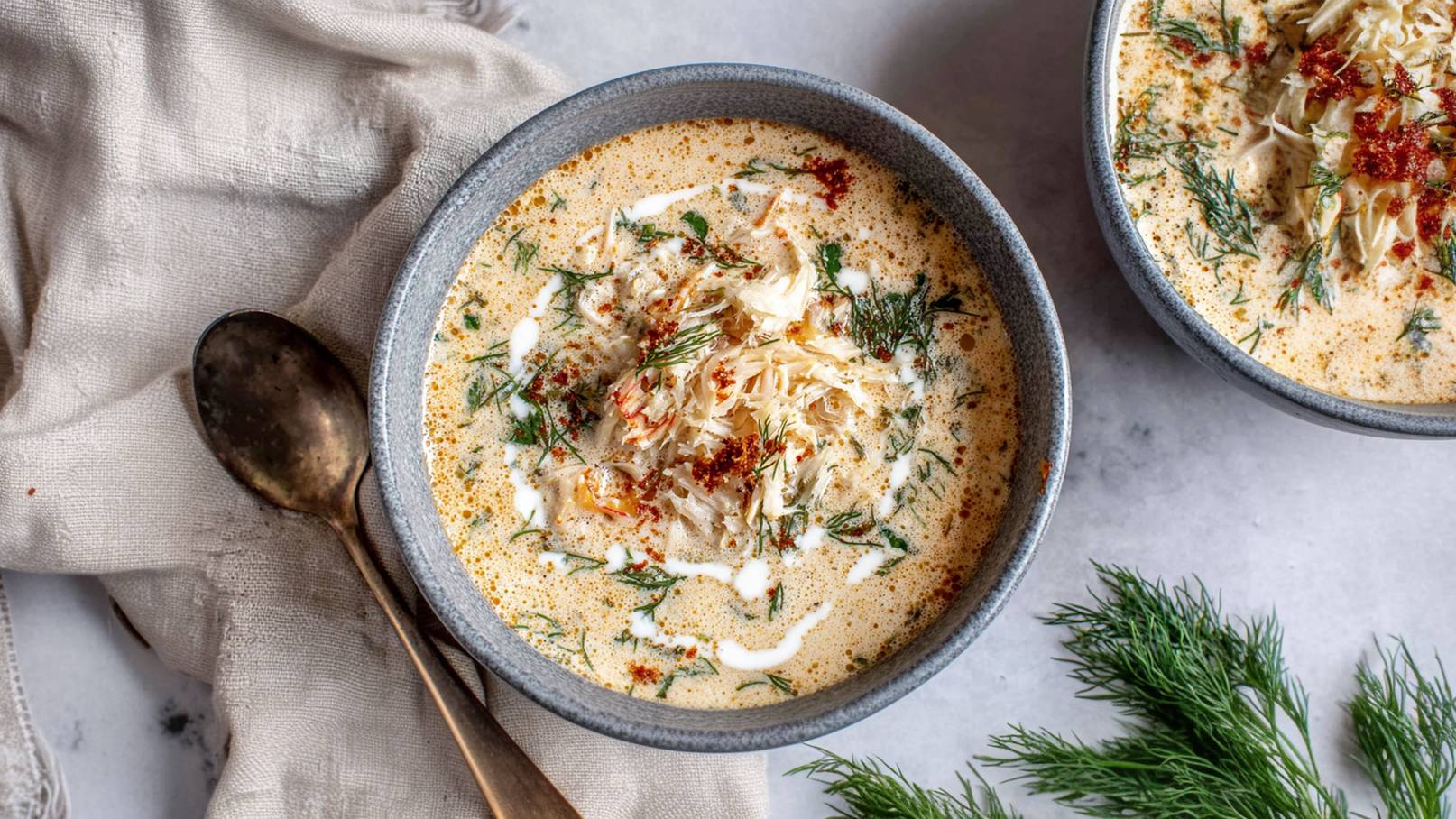 Eine herzliche Suppe in einer grauen Schüssel, garniert mit frischem Dill, Sahne und einer Prise Paprika. Der Rand der Schüssel zeigt eine cremige, goldene Flüssigkeit. Im Hintergrund sind weitere Schalen sichtbar, umgeben von einem gedeckten Tisch.