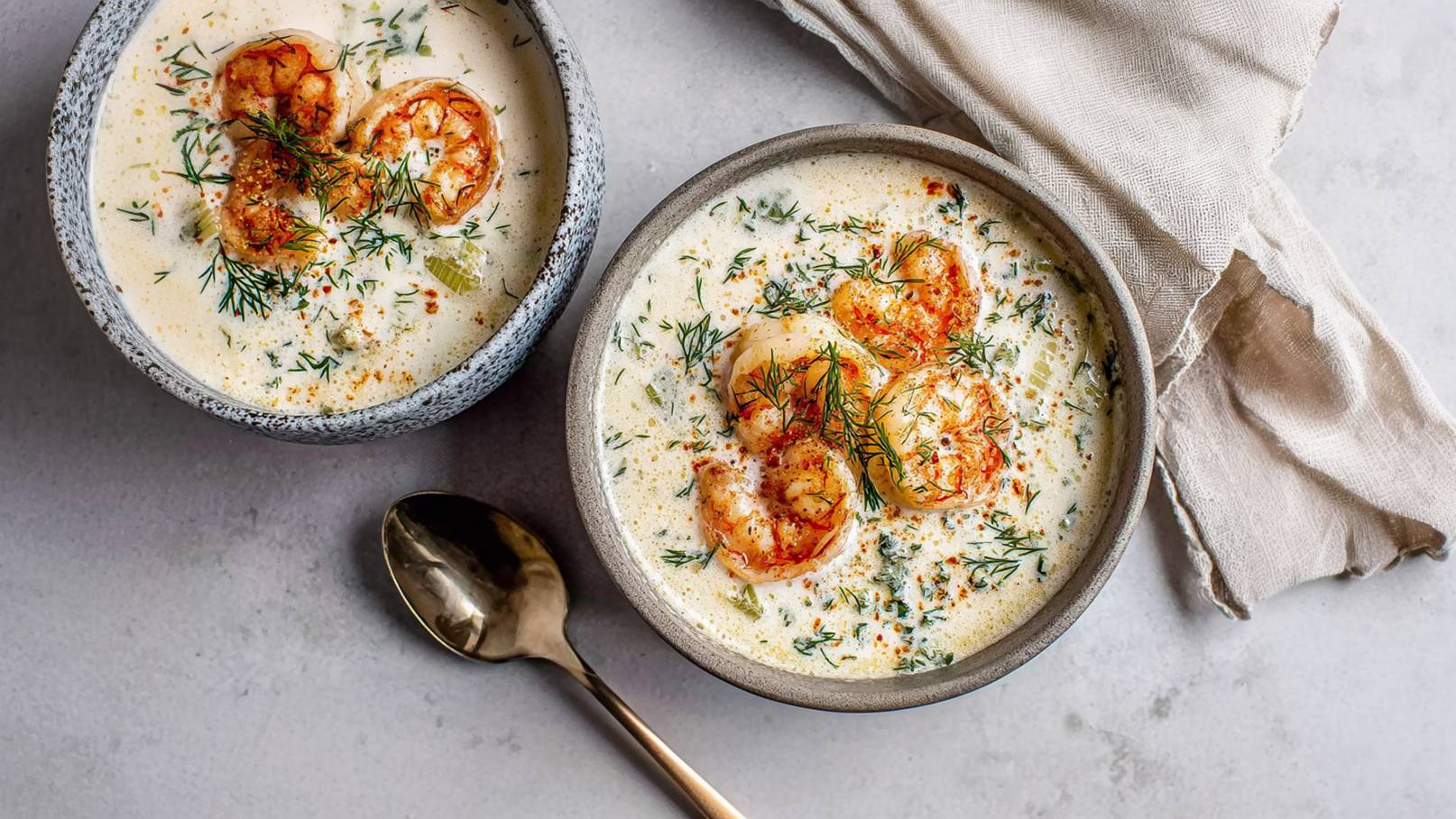 Zwei Schalen mit cremiger Garnelensuppe sind auf einer Tischplatte angeordnet. Die Suppe enthält mehrere große Garnelen, frischen Dill und Gewürze. Ein Löffel liegt neben den Schalen. Ein feuchtes Tuch ist ebenfalls sichtbar.