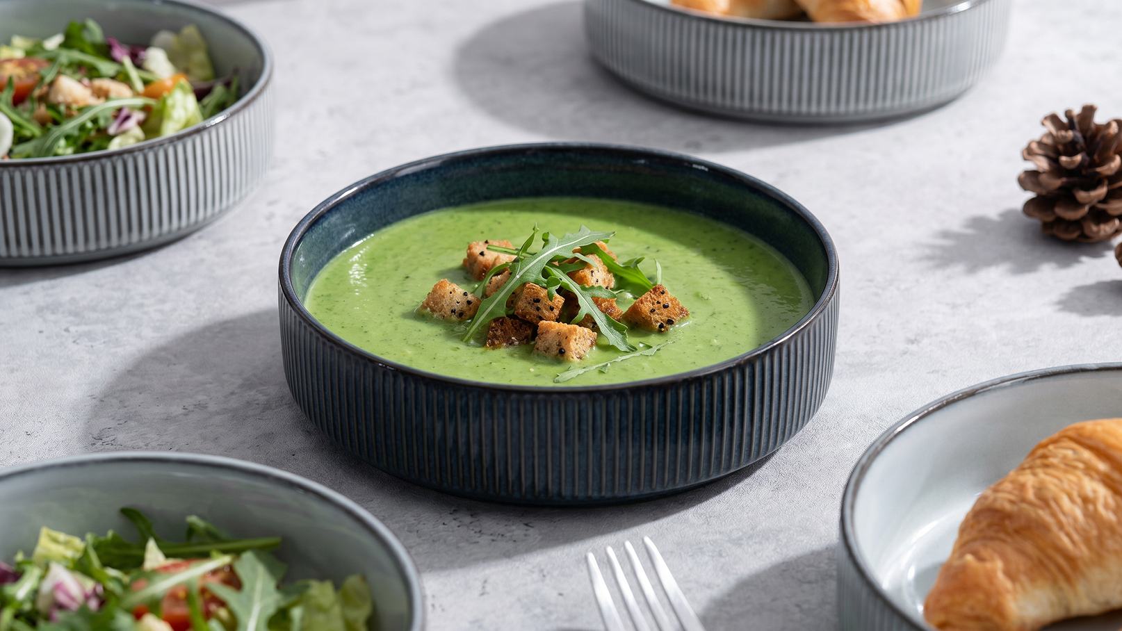 Eine grüne Suppe in einer runden, tiefen Schüssel, garniert mit Croutons und frischen Kräutern. Um die Suppe sind mehrere Schüsseln mit Salat und ein Croissant angeordnet, auf einem grauen Tisch.