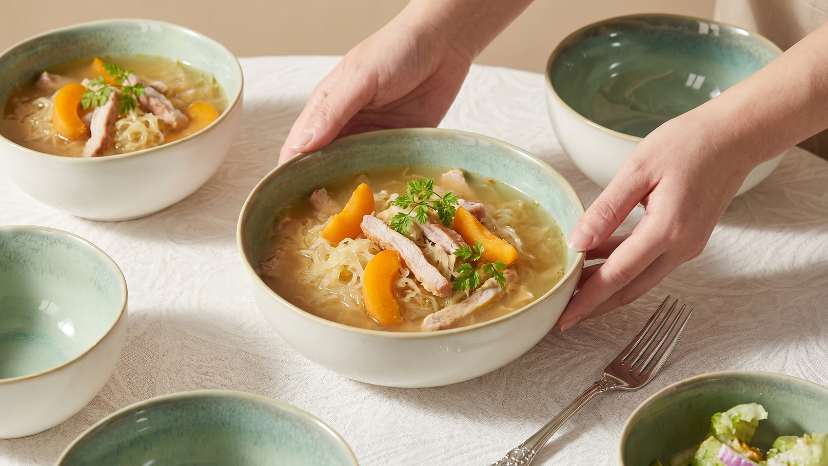 Eine Person reicht eine Schüssel mit einer klaren Brühe, daneben sind weitere leere Schalen. In der Brühe befinden sich Nudeln, Stücke von Fleisch und orangefarbene Möhrenscheiben. Der Tisch ist mit einer hellen Tischdecke gedeckt.