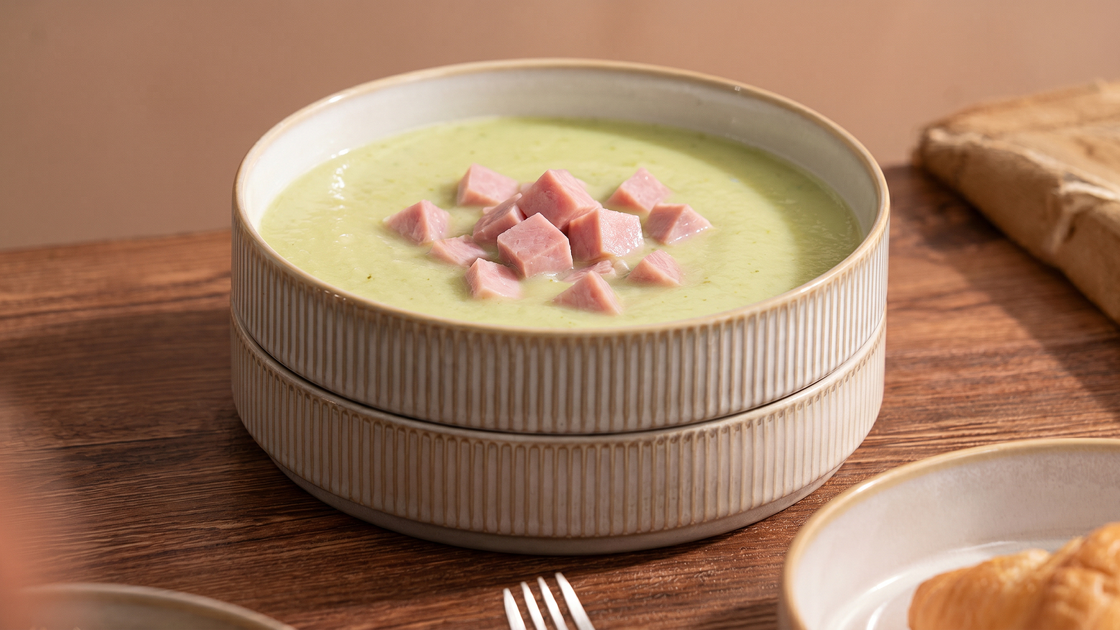 Eine cremige grüne Suppe in einer doppelten Schüssel, garniert mit kleinen Würfeln von Schinken. Der Hintergrund zeigt eine Holzoberfläche. Zubehör wie Besteck und ein Croissant sind ebenfalls sichtbar.