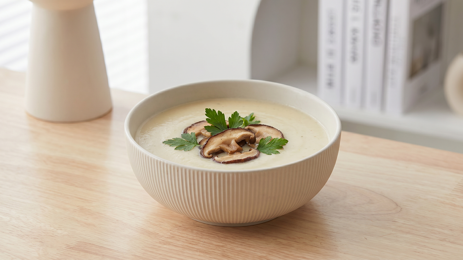 Eine schlichte, weiße Schüssel mit cremiger Suppe steht auf einem Holztisch. Die Suppe ist mit einigen Scheiben Pilzen und frischer Petersilie garniert. Im Hintergrund sind verschwommen Bücher und eine Vase sichtbar.