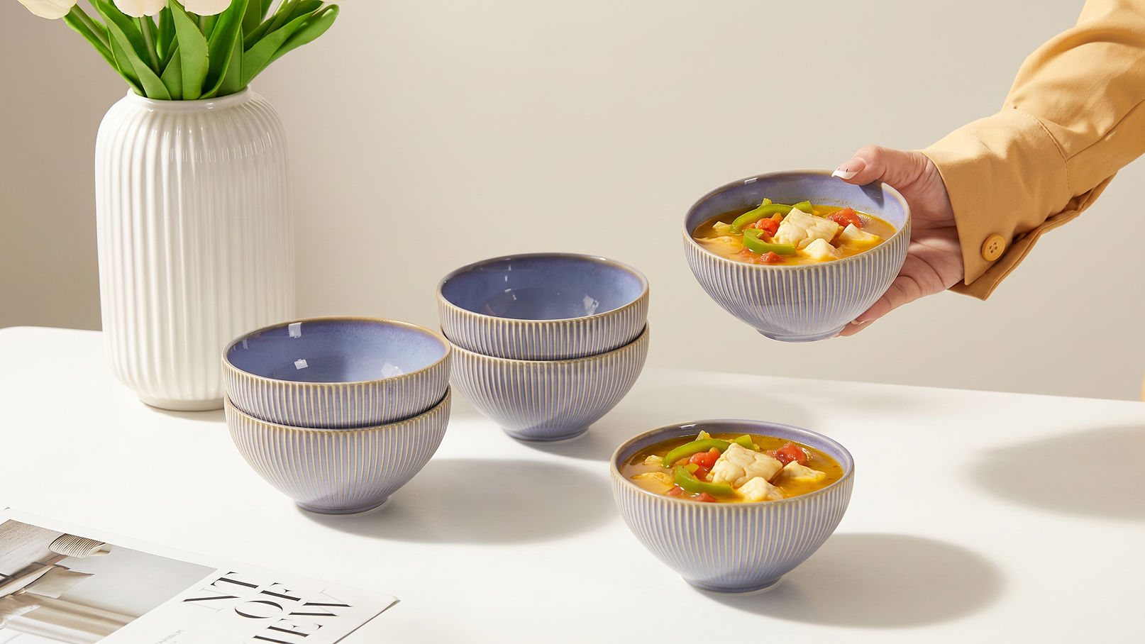 Eine Hand reicht eine Schüssel Suppe mit Gemüse und Hähnchenstücken. Auf dem Tisch stehen weitere Schüsseln in verschiedenen Größen mit einem eleganten blauen Rand. Im Hintergrund sieht man eine Vase mit frischen Blumen.