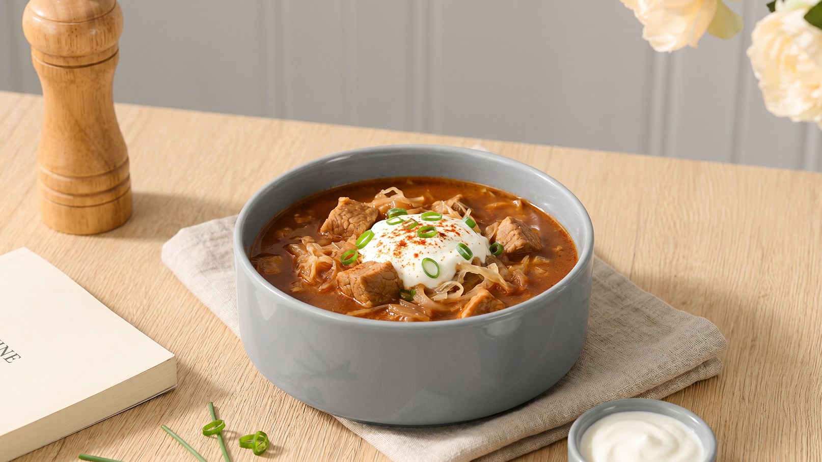 Eine graue Schüssel enthält eine herzhafte Suppe mit Fleischstücken, Gemüse und einer großen Portion Sahne oder Joghurt oben drauf. Die Suppe ist mit frischen Frühlingszwiebeln und roten Chiliflocken garniert. Ein Holzstreuer und ein Buch liegen neben der Schüssel auf einem Tisch.