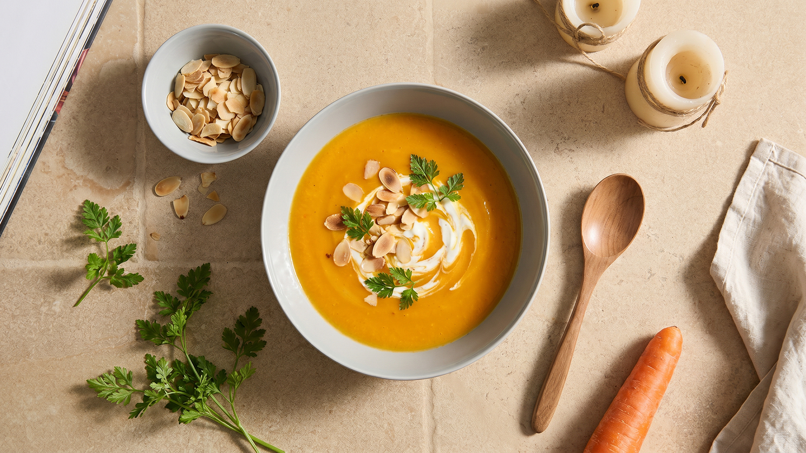 Eine Schüssel mit cremiger Karottensuppe, garniert mit Sahne, Mandelscheiben und frischem Koriander. Neben der Schüssel steht eine kleine Schale mit Mandeln. Im Hintergrund sind ein Karotte und einige Kerzen zu sehen. Der Tisch hat eine neutrale, sandfarbene Oberfläche.