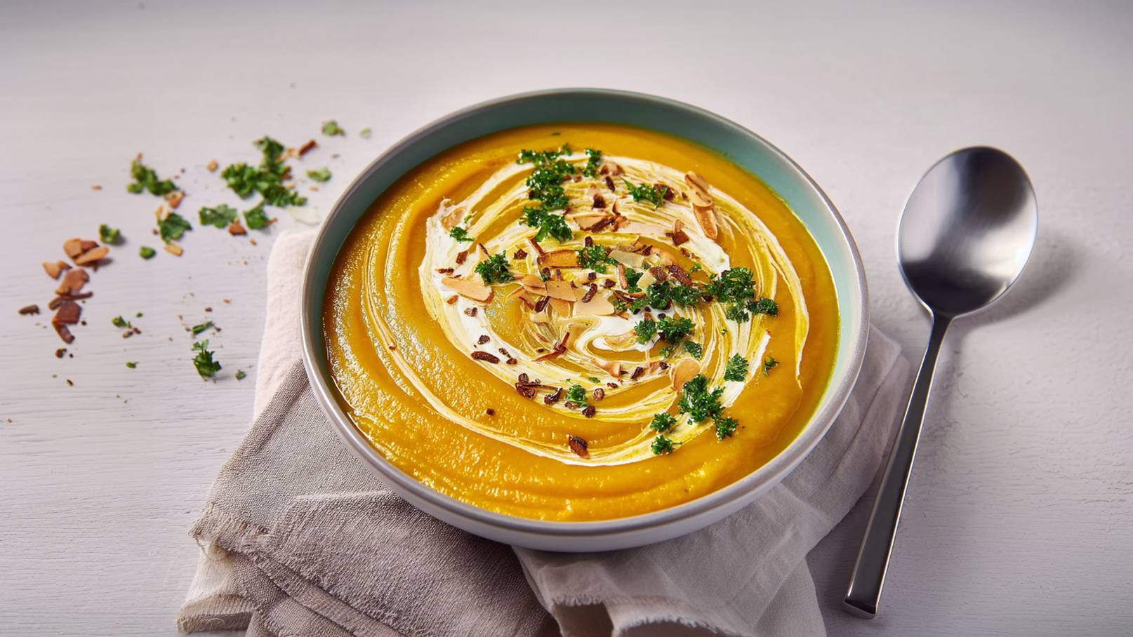 Eine Schüssel mit cremiger Kürbissuppe, garniert mit Sahne, gerösteten Mandeln und frischer Petersilie. Die Suppe hat eine leuchtend orange Farbe und liegt auf einem hellen Untergrund. Daneben liegt ein Löffel und ein Stofftuch.