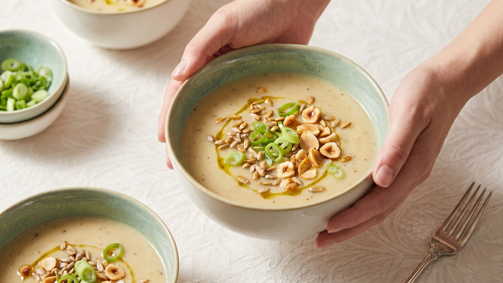 Eine Person hält eine Schüssel mit cremiger Suppe, die mit gerösteten Nüssen und grünen Zwiebeln garniert ist. Im Hintergrund befinden sich weitere Schüsseln mit ähnlicher Suppe und eine kleine Schale mit geschnittenen Zwiebeln. Der Tisch ist mit einem weißen Tuch bedeckt.