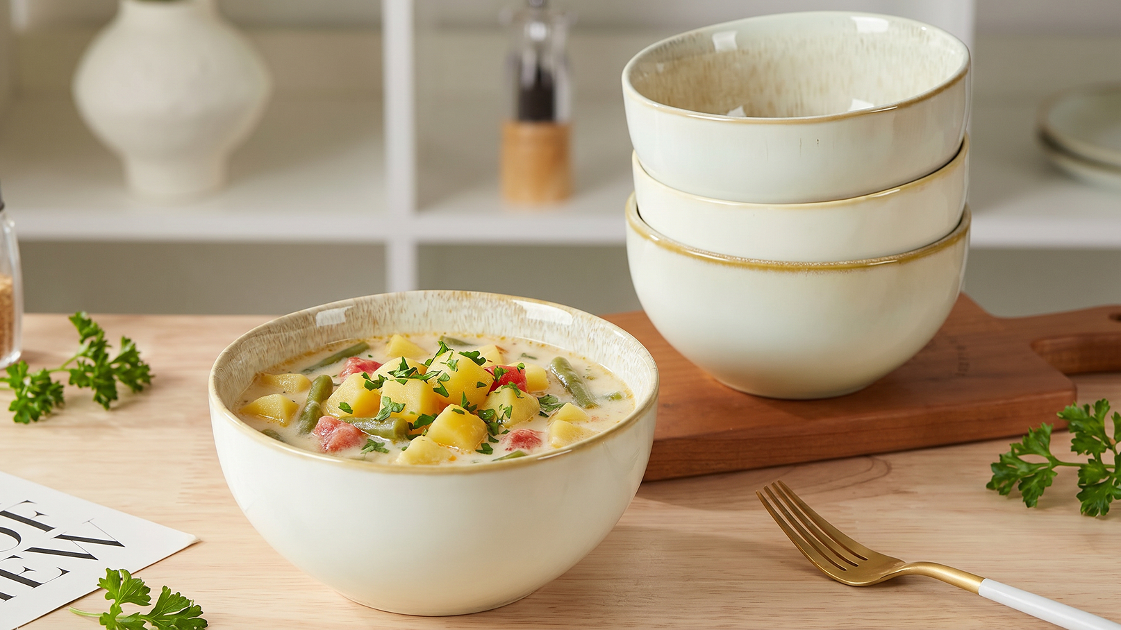Eine weiße Schüssel mit einer bunten Suppe, die Kartoffeln, Gemüse und Kräuter enthält, steht auf einem Holzbrett. Im Hintergrund sind drei stapelbare, ähnliche Schüsseln zu sehen. Einige Kräuterblätter liegen auf dem Tisch.