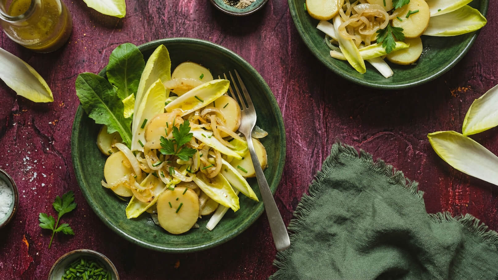 Zwei grüne Teller mit einem Salat aus gekochten Kartoffeln, Zwiebeln und Endiviensalat, garniert mit frischer Petersilie und Schnittlauch. Neben den Tellern befinden sich kleine Schalen mit Salz und Gewürzen sowie ein grünes Tuch. Der Hintergrund ist strukturiert und in dunklen Farben gehalten.
