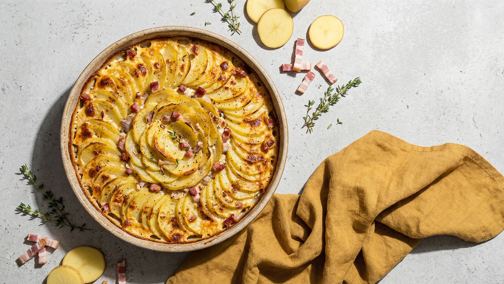 Ein runder Auflauf mit geschichteten Kartoffelscheiben, garniert mit Speckwürfeln und frischen Kräutern. Der goldbraun gebackene Auflauf liegt in einer flachen Schüssel auf einer neutralen Oberfläche, umgeben von rohen Kartoffelscheiben und einer gelben Serviette.