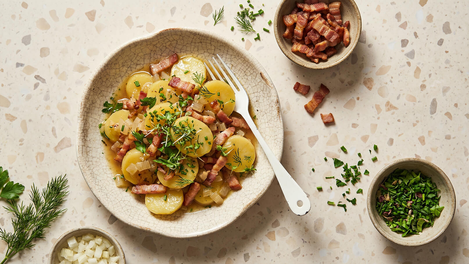 Ein Teller mit Kartoffelsalat, garniert mit Speck, Zwiebeln und frischen Kräutern wie Dill und Schnittlauch. Eine Gabel liegt neben dem Teller. Weitere kleine Schalen mit gehacktem Speck und frischen Kräutern sind in der Nähe platziert. Der Hintergrund ist neutral.