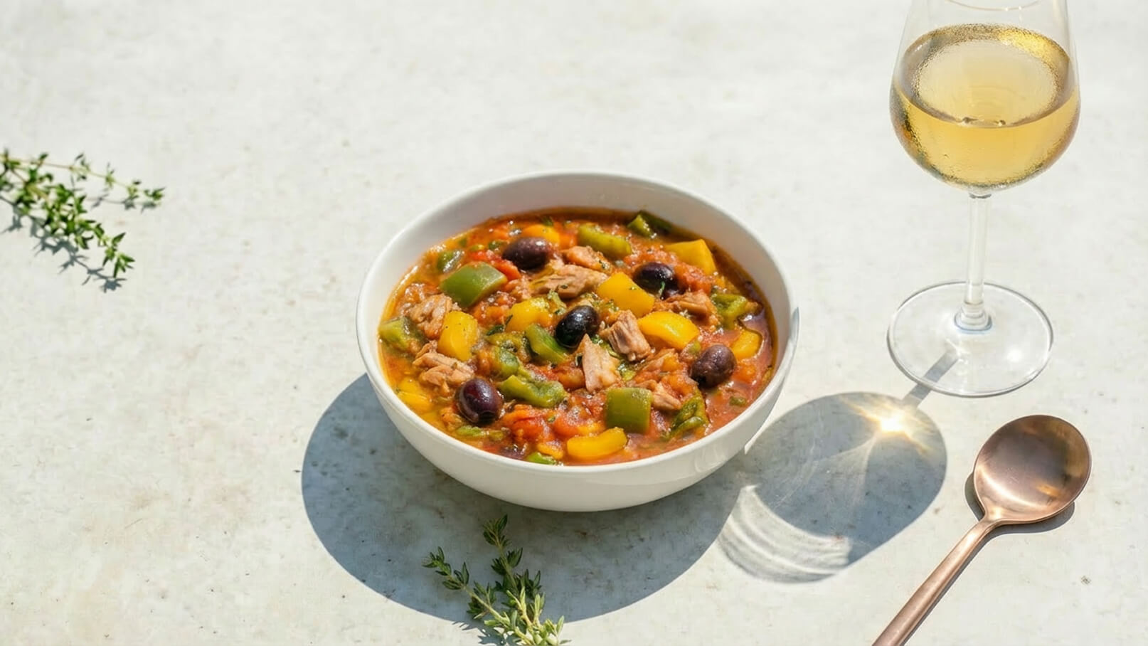 Eine Schüssel mit buntem Eintopf, der Stücke von Fleisch, Paprika und Oliven enthält. Daneben steht ein Glas Weißwein. Das Gericht präsentiert sich in einem hellen, einladenden Ambiente.