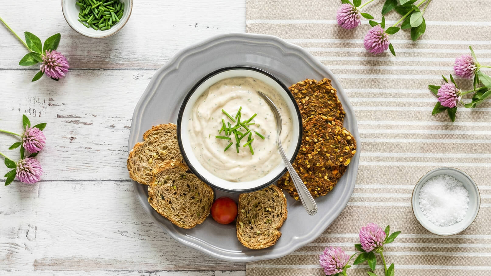Ein grauer Teller mit einem Schälchen Dip, garniert mit Schnittlauch, und umgeben von verschiedenen Brotscheiben und einer kleinen Tomate. Neben dem Teller befindet sich eine Schüssel mit Salz. Die Anordnung ist auf einem hellen, gestreiften Tischtuch platziert.