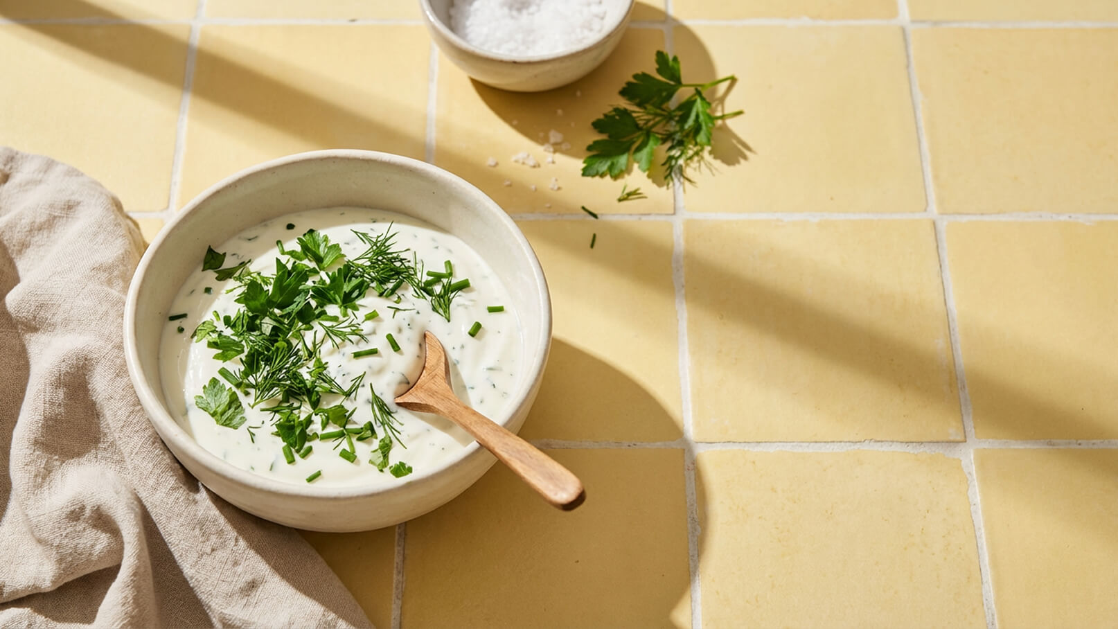 Eine Schale mit cremigem Dip, garniert mit frischen Kräutern wie Schnittlauch und Dill. Ein Holzlöffel liegt in der Schale. Im Hintergrund sind ein kleine Schüssel mit Salz und frische Petersilie zu sehen. Der Untergrund ist gelb gefliest.