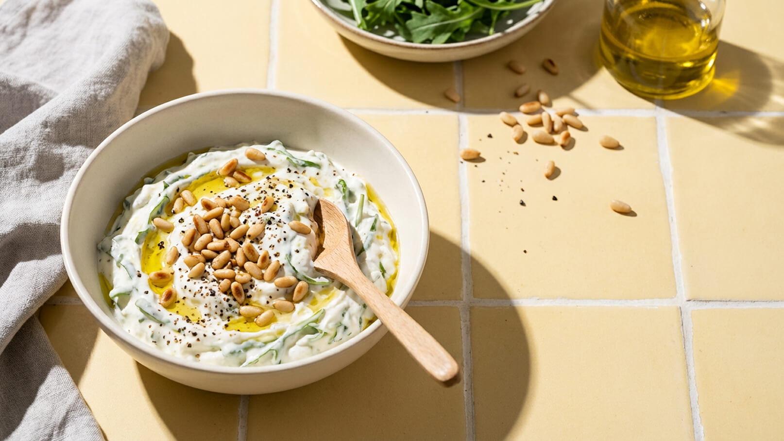 Eine Schüssel mit cremigem Dip, garniert mit Pinienkernen und einem Spritzer Olivenöl. Daneben liegt ein Holzlöffel und einige schwarze Pfefferkörner sind verstreut. Im Hintergrund befindet sich eine Schüssel mit frischem Blattspinat.