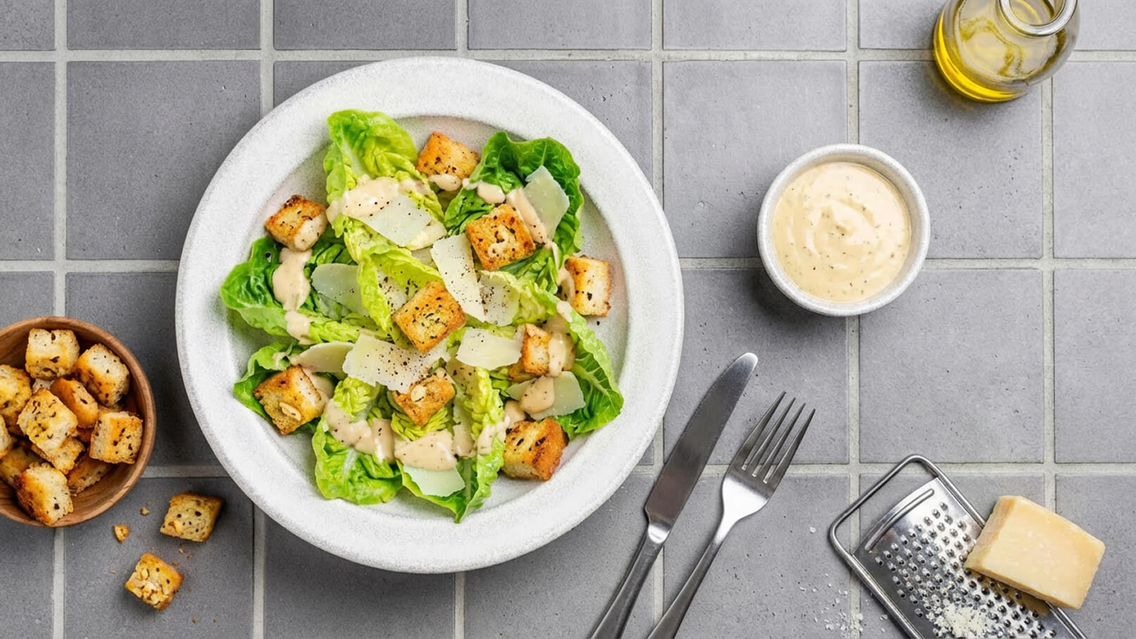 Ein Teller mit Caesar-Salat, garniert mit Römersalat, Croutons, Parmesan und einem cremigen Dressing. Neben dem Teller steht eine Schüssel mit zusätzlichem Dressing und eine Handvoll gerösteter Croutons auf einem grauen Fliesenboden.
