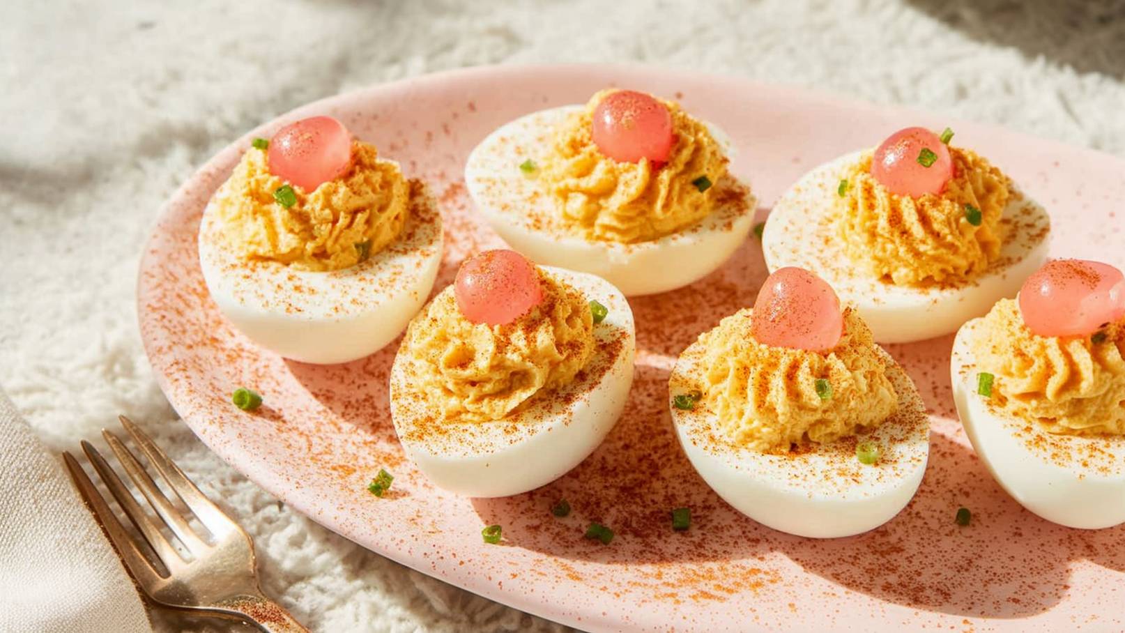 Auf einem rosa Teller liegen sechs halbe Eier, gefüllt mit einer gelben Creme und garniert mit kleinen, roten Gelee-Kugeln sowie etwas Paprika. Frische Kräuter sind ebenfalls sichtbar. Die Eier sind farbenfroh angerichtet und wirken appetitlich.