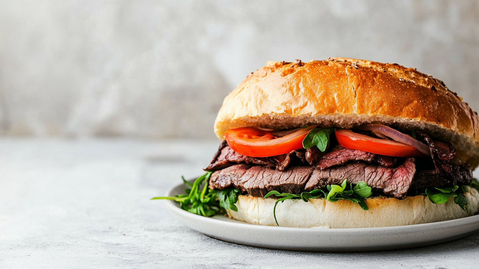 Ein frisch gebratener Sandwich mit saftigem Rindfleisch, frischen Tomatenscheiben, Zwiebeln und Kräutern. Das Brötchen ist knusprig und goldbraun, serviert auf einem Teller mit einer Unterlage aus Rucola.