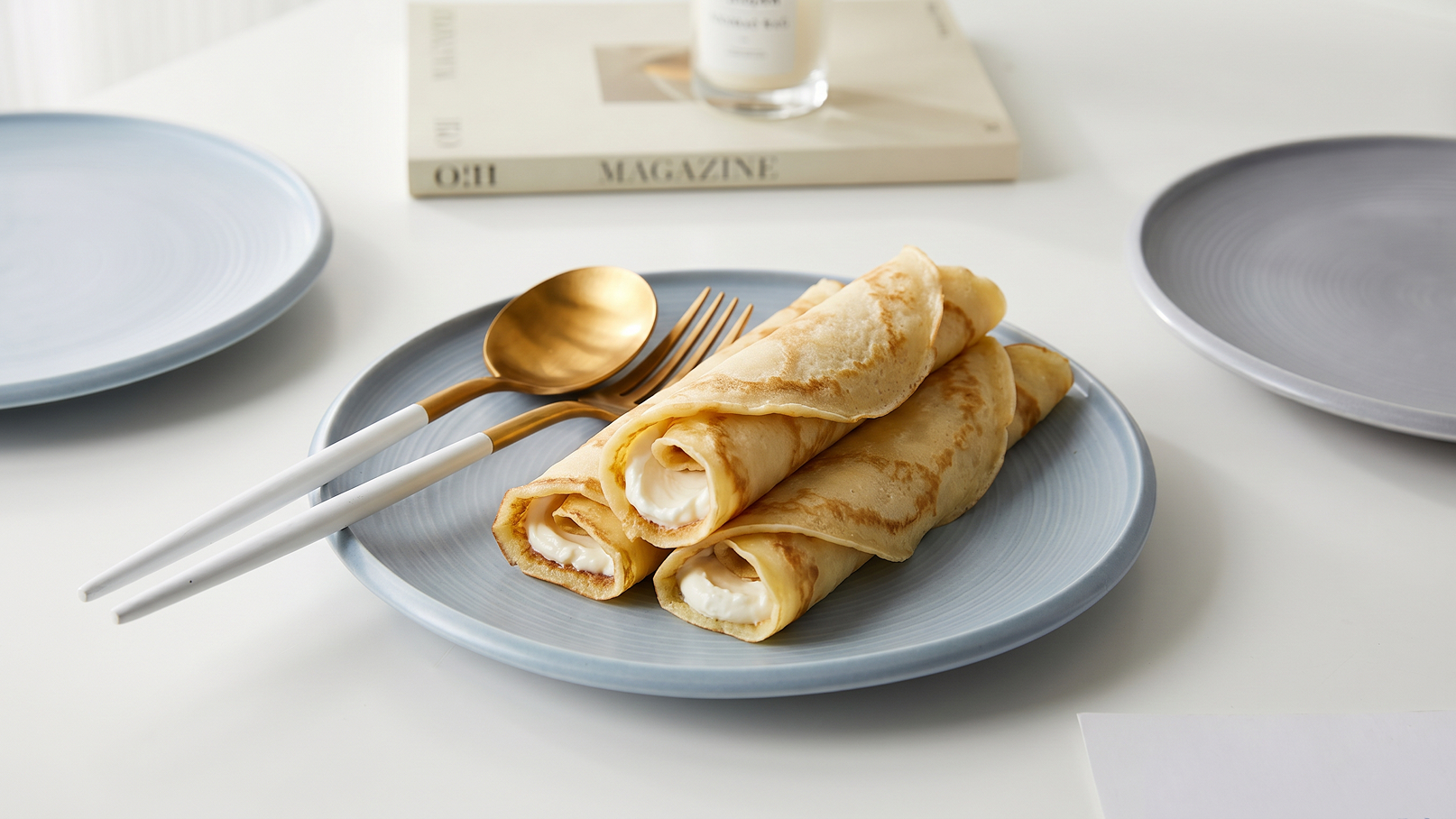 Auf einem blauen Teller liegen mehrere aufgerollte Pfannkuchen, gefüllt mit einer weißen Creme. Daneben liegen eine goldene Gabel und ein Löffel sowie zwei graue Teller im Hintergrund. Ein Buch und eine Kerze sind ebenfalls zu sehen, jedoch weniger auffällig.