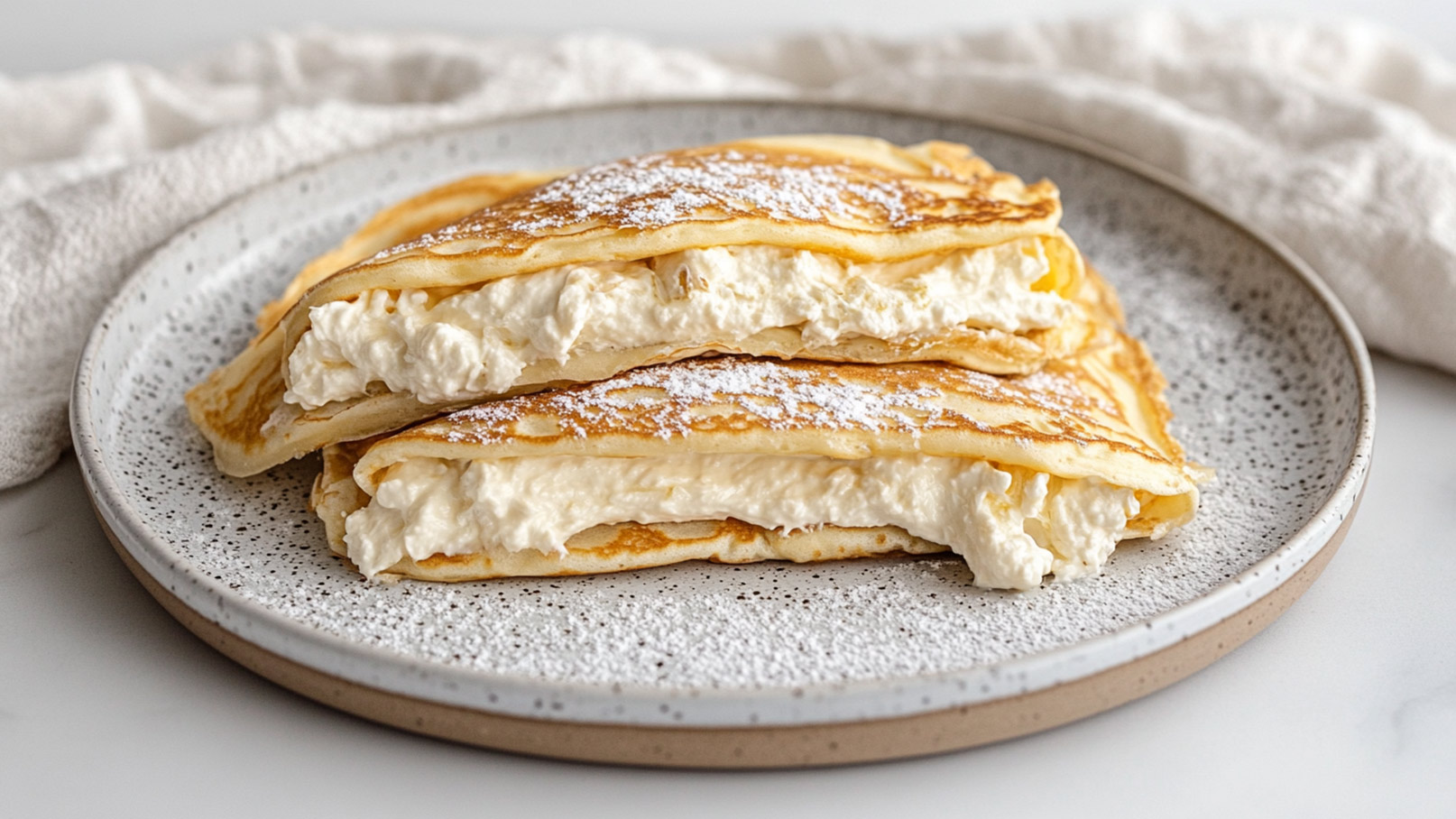 Ein Teller mit zwei übereinander liegenden Pfannkuchen, gefüllt mit einer cremigen Füllung. Die Oberseite der Pfannkuchen ist leicht mit Puderzucker bestäubt. Der Teller hat einen schlichten, grauen Rand und ist auf einem hellen Untergrund platziert.