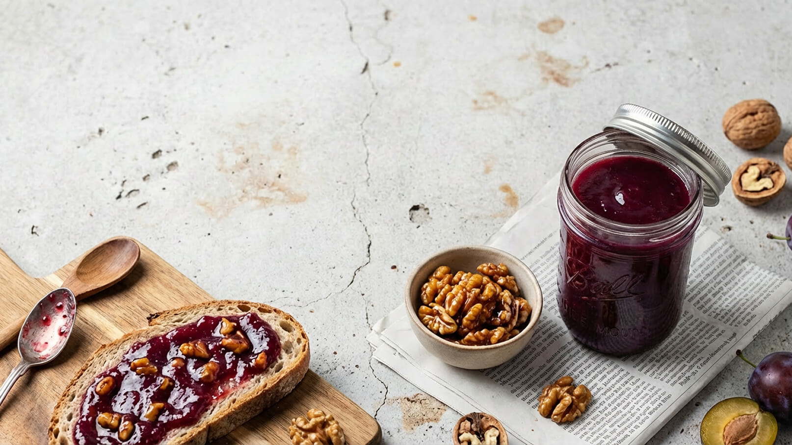 Ein Stück Vollkornbrot mit roter Marmelade und gehackten Walnüssen liegt auf einem Holzbrett. Daneben steht ein offenes Glas mit Marmelade und eine Schüssel mit Walnüssen. Der Hintergrund ist neutral und strahlend.