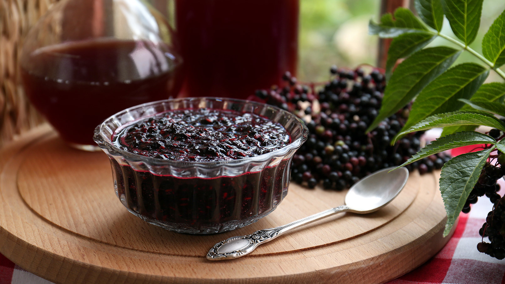 Ein Glas mit dunkler Holunderkonfitüre steht auf einem Holzbrett. Daneben liegt ein silberner Löffel. Im Hintergrund befinden sich Holunderbeeren und ein Krug mit Holundergetränk. Die Szene vermittelt einen heimeligen, naturnahen Eindruck.