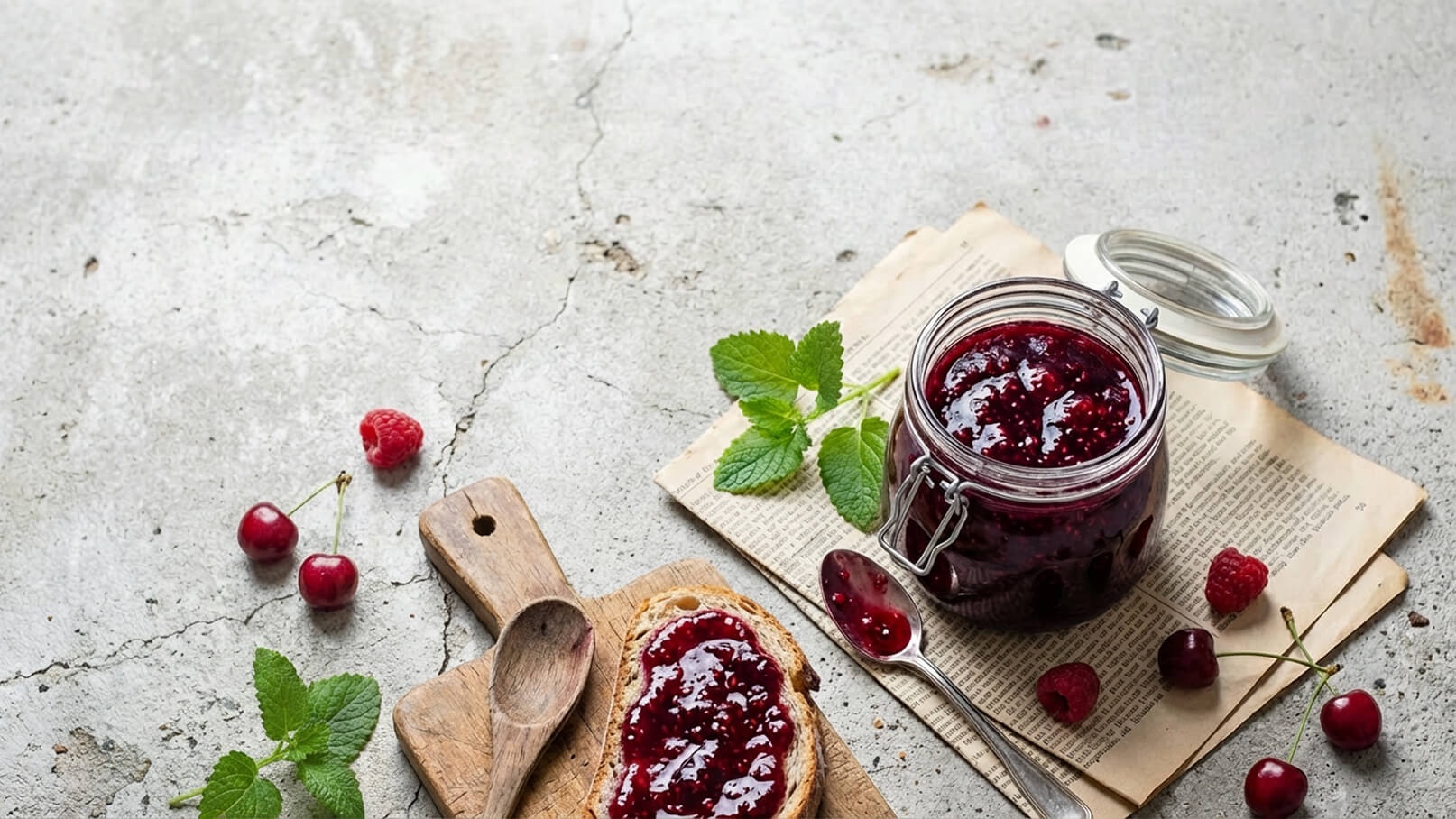 Ein Glas Obstkonfitüre steht auf einer offenen Seite eines Buches. Daneben liegt ein Stück Brot, das mit der Konfitüre bestrichen ist. Einige frische Himbeeren und Kirschen sowie ein Holzlöffel liegen ebenfalls in der Nähe.