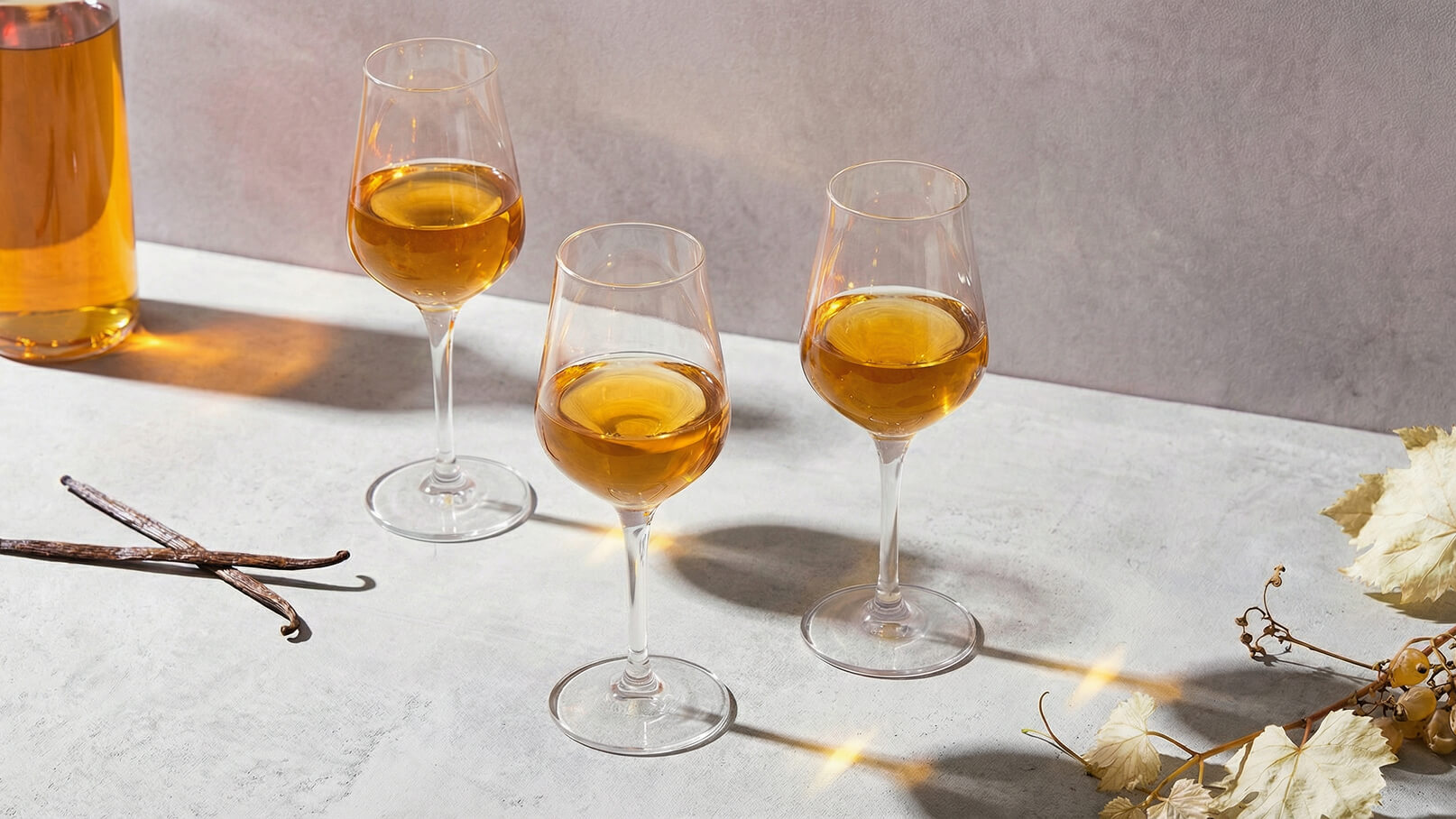 Drei Weingläser mit goldenem Getränk stehen auf einem hellen Tisch. Neben den Gläsern liegt eine Flasche mit demselben Getränk. Einige Vanilleschoten sind ebenfalls auf dem Tisch platziert, was eine einladende und stilvolle Atmosphäre schafft.